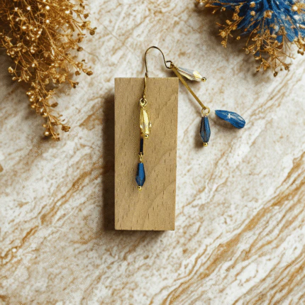 Boucles d'oreilles longues graphiques dorées, cristal transparent et goutte bleue, accrochées à un bloc de bois