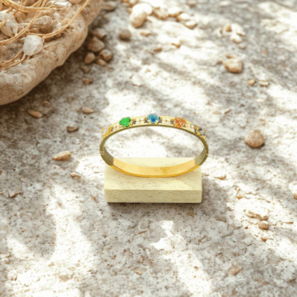 Bracelet jonc arc-en-ciel doré posé sur un petit socle en bois, galets et sable en fond