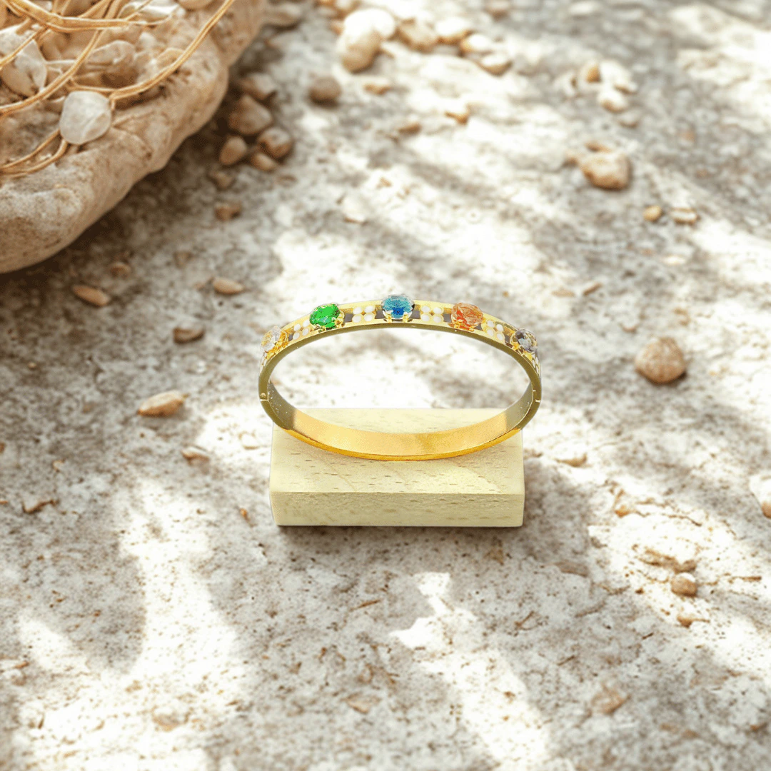 Bracelet jonc arc-en-ciel doré posé sur un petit socle en bois, galets et sable en fond