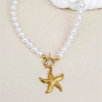 Collier de perles blanches à fermoir T et pendentif étoile de mer doré posé à plat près d'un coquillage