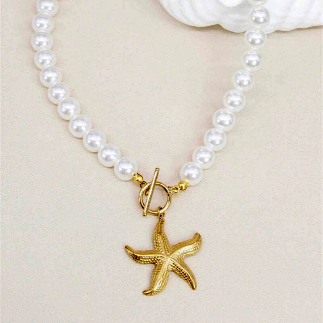 Collier de perles blanches à fermoir T et pendentif étoile de mer doré posé à plat près d'un coquillage