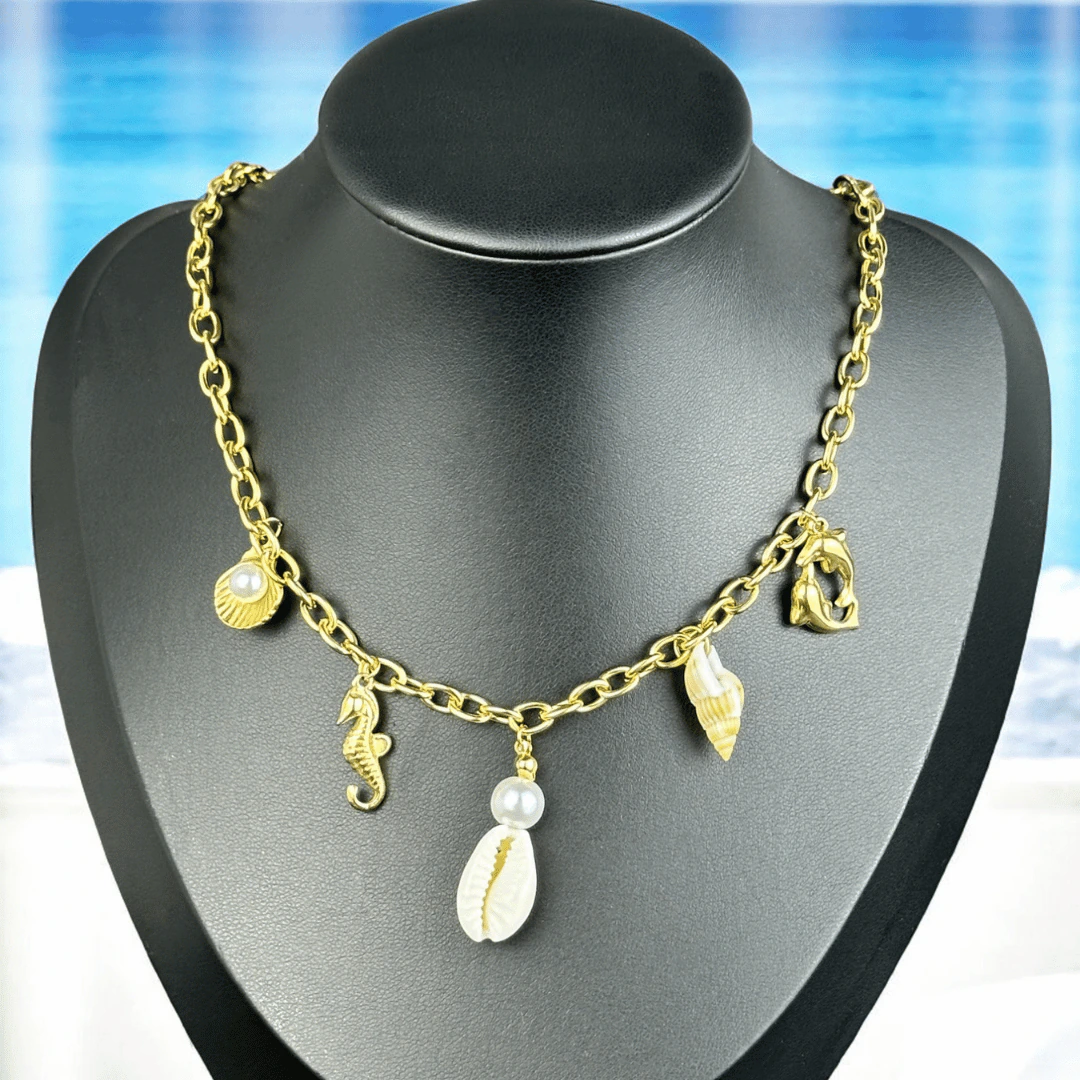 Collier chaîne dorée à breloques marines, coquillages et perles sur buste noir, fond de plage