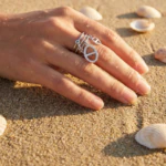 Bague femme argentée ouverte à charms ovales sertis de strass portée à la main sur le sable