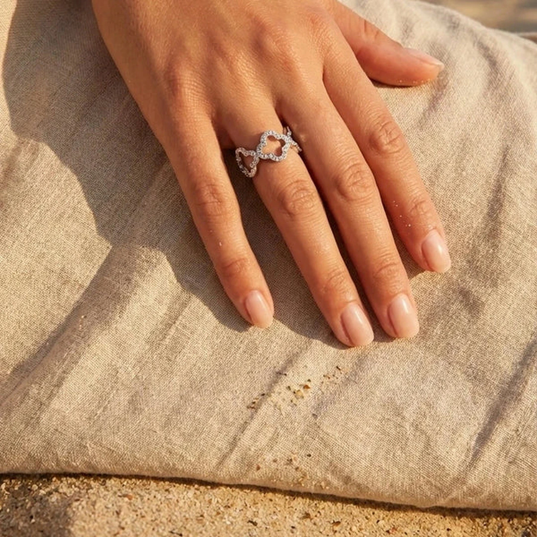 Main portant une bague argentée à trèfles ajourés pavés de strass, posée sur un tissu en lin