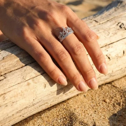 Bague femme argentée motif couronne sertie de strass portée au doigt, main posée sur du bois flotté