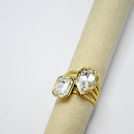 Bague femme dorée à pierres facettées transparentes rectangle et goutte, anneau multirang sur support beige