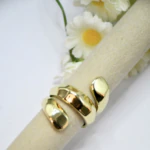 Bague ouverte dorée effet ruban enroulé sur un support beige devant des marguerites blanches