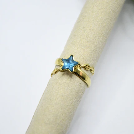 Bague dorée à étoile bleue facettée, anneau lisse et chaîne fine superposés sur un support beige