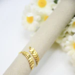 Bague ouverte dorée à branches de feuilles croisées posée sur un support beige près de marguerites