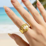 Bague femme dorée motif fleur texturée au centre façon perle blanche portée à la main devant la mer