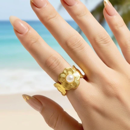 Bague femme dorée motif fleur texturée au centre façon perle blanche portée à la main devant la mer