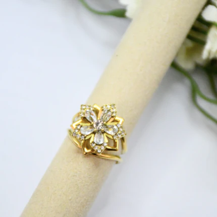 Bague fleur dorée aux pierres transparentes posée sur un rouleau beige, vue large avec fleurs blanches