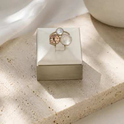 Bague dorée trois pierres effet nacré et rosé sur un présentoir cube blanc posé sur une pierre beige