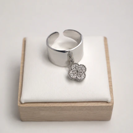 Bague ouverte argentée à anneau large et lisse avec charm trèfle suspendu sur un présentoir blanc
