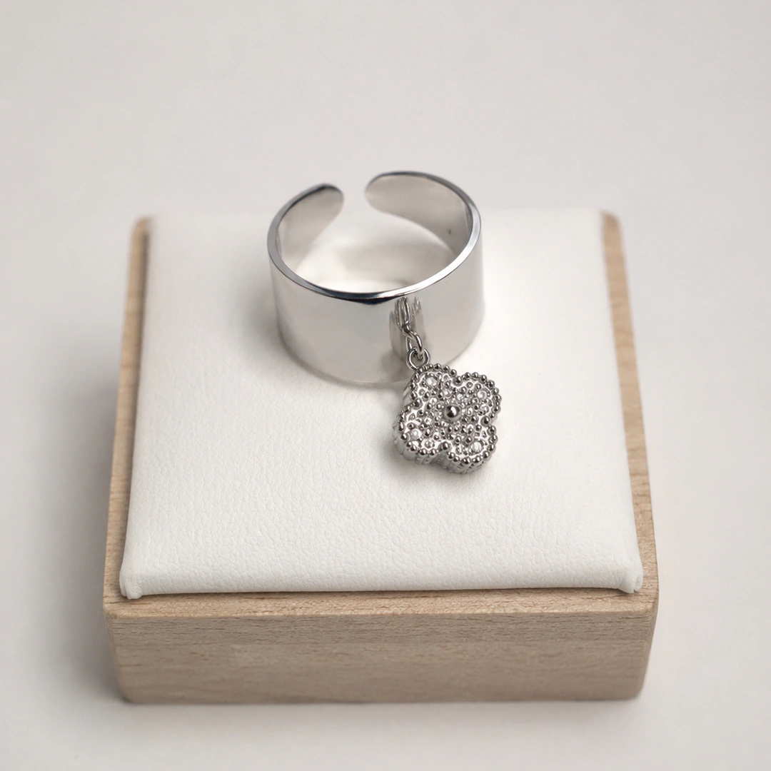 Bague ouverte argentée à anneau large et lisse avec charm trèfle suspendu sur un présentoir blanc