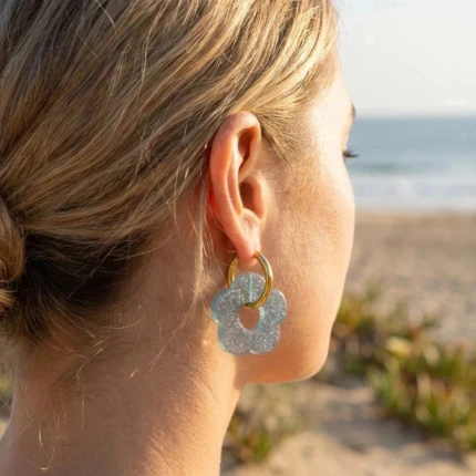 Boucles d'oreilles créoles dorées à fleur bleue pailletée portées à l'oreille face à la mer
