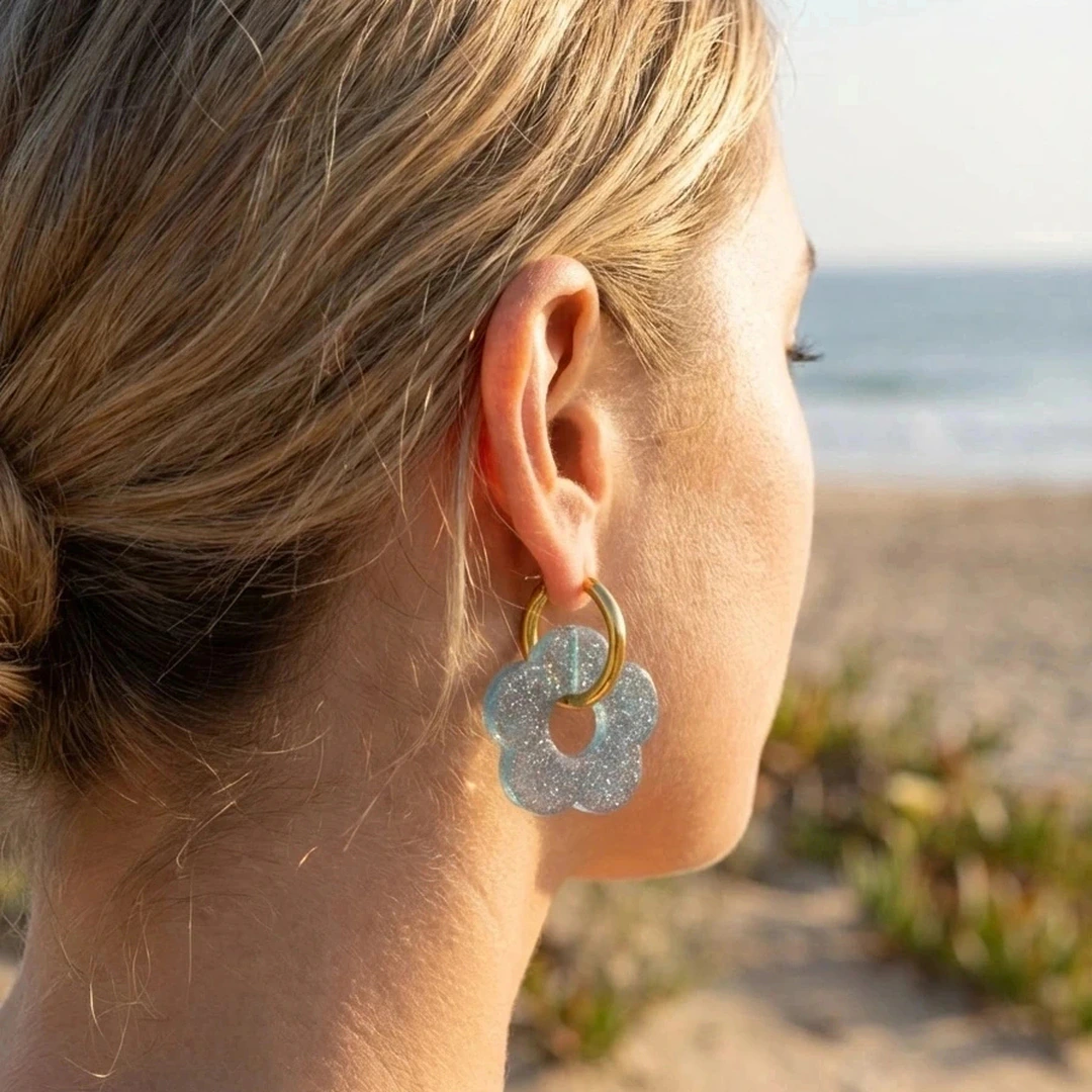 Boucles d'oreilles créoles dorées à fleur bleue pailletée portées à l'oreille face à la mer
