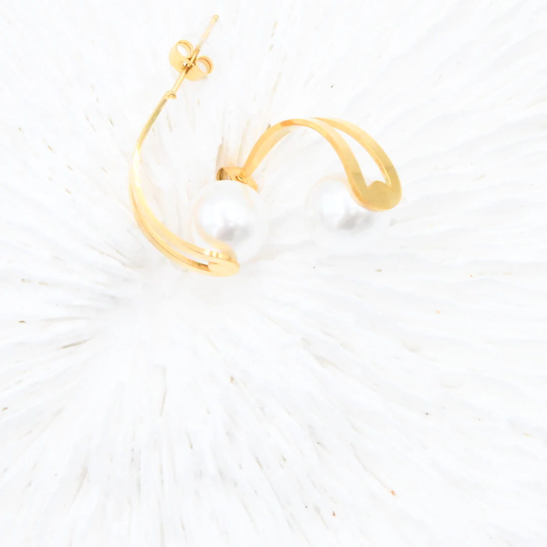 Paire de boucles d'oreilles dorées courbées à perle blanche posée sur un fond blanc duveteux