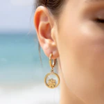Boucles d'oreilles créoles dorées médaillon fleur sur fond blanc nacré portées à l'oreille, mer en fond