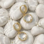 Paire de créoles dorées à pendentif rond transparent posée sur des galets blancs mouillés