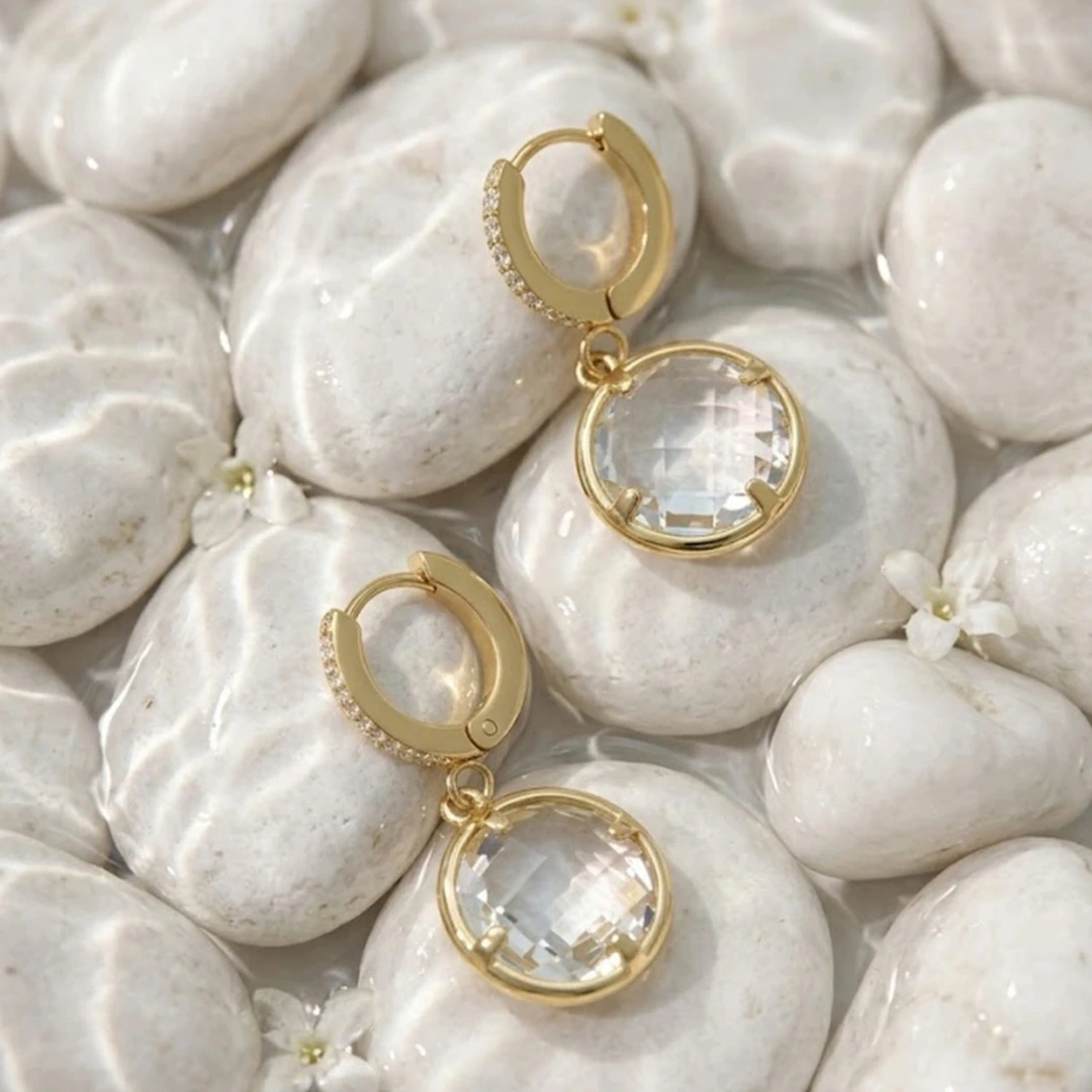 Paire de créoles dorées à pendentif rond transparent posée sur des galets blancs mouillés