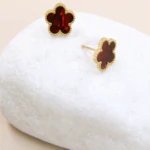 Boucles d'oreilles clous fleur rouge dorées au contour perlé posées sur un galet blanc
