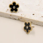 Clous d'oreilles fleur noire à bordure dorée perlée et cristal central, l'un posé sur une pierre beige