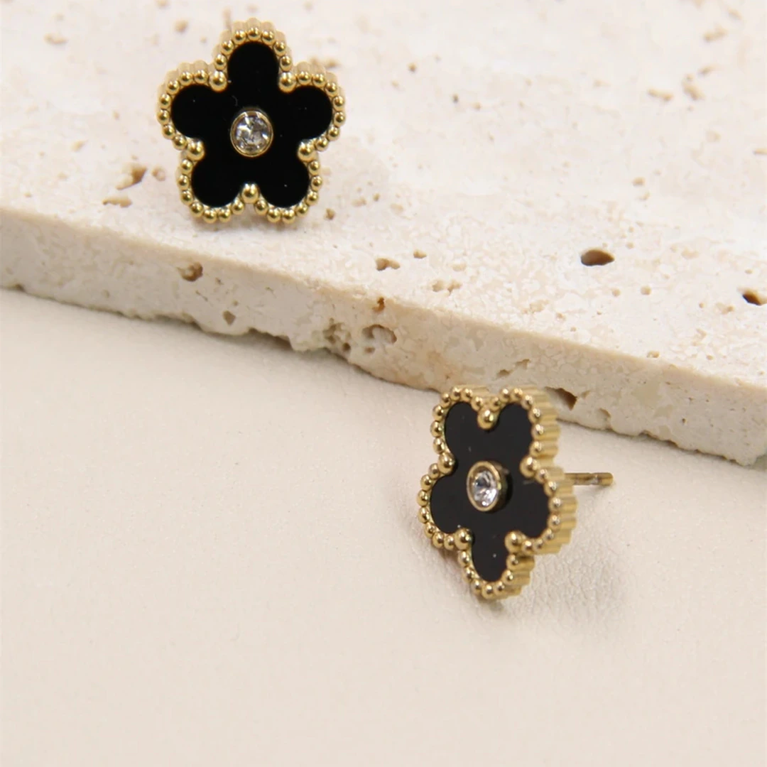 Clous d'oreilles fleur noire à bordure dorée perlée et cristal central, l'un posé sur une pierre beige