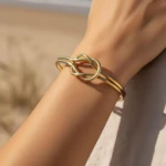 Bracelet jonc doré ouvert femme à motif nœud entrelacé porté au poignet, plage en arrière-plan