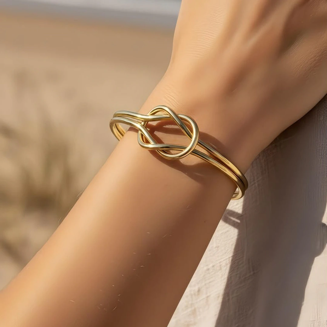 Bracelet jonc doré ouvert femme à motif nœud entrelacé porté au poignet, plage en arrière-plan