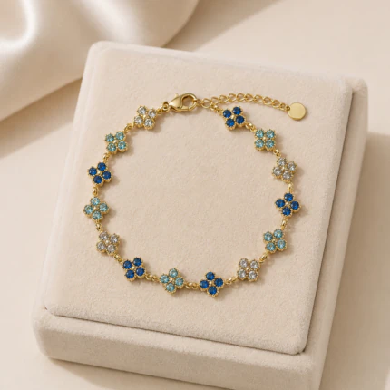 Bracelet doré femme à petits trèfles scintillants en strass bleu nuit, bleu clair et blanc, posé sur un écrin