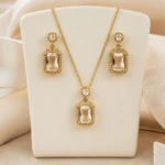 Collier et boucles d'oreilles dorés à pendentifs rectangulaires cerclés de strass sur présentoir crème