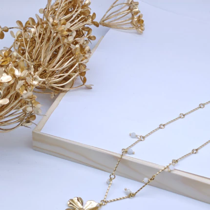 Collier chaîne fine à pendentif fleur dorée et perles blanches posé près de fleurs séchées dorées