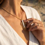 Collier doré femme à pendentif feuille stylisée et détail blanc, porté au cou et tenu entre les doigts