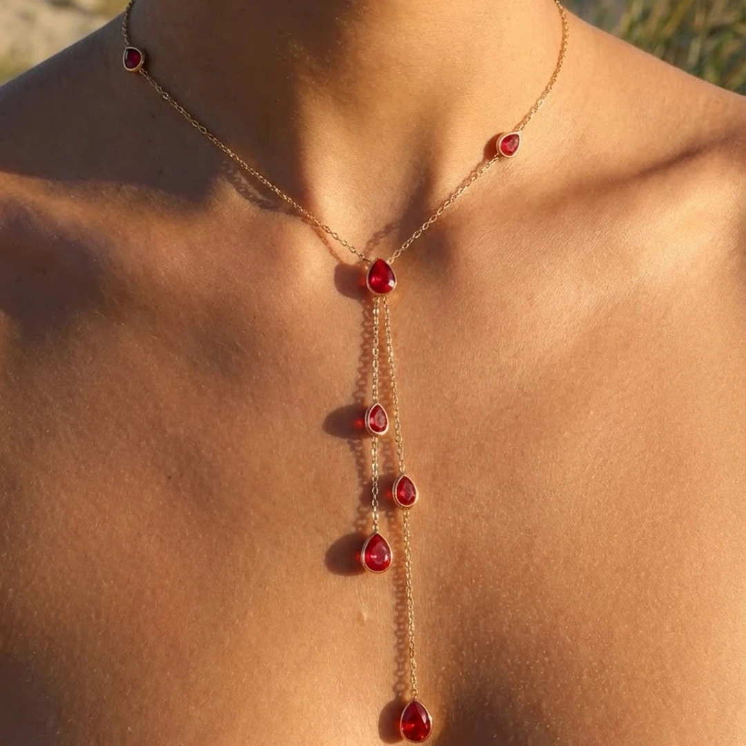 Collier doré femme en Y à gouttes rouges pendantes porté au cou, vue rapprochée au soleil