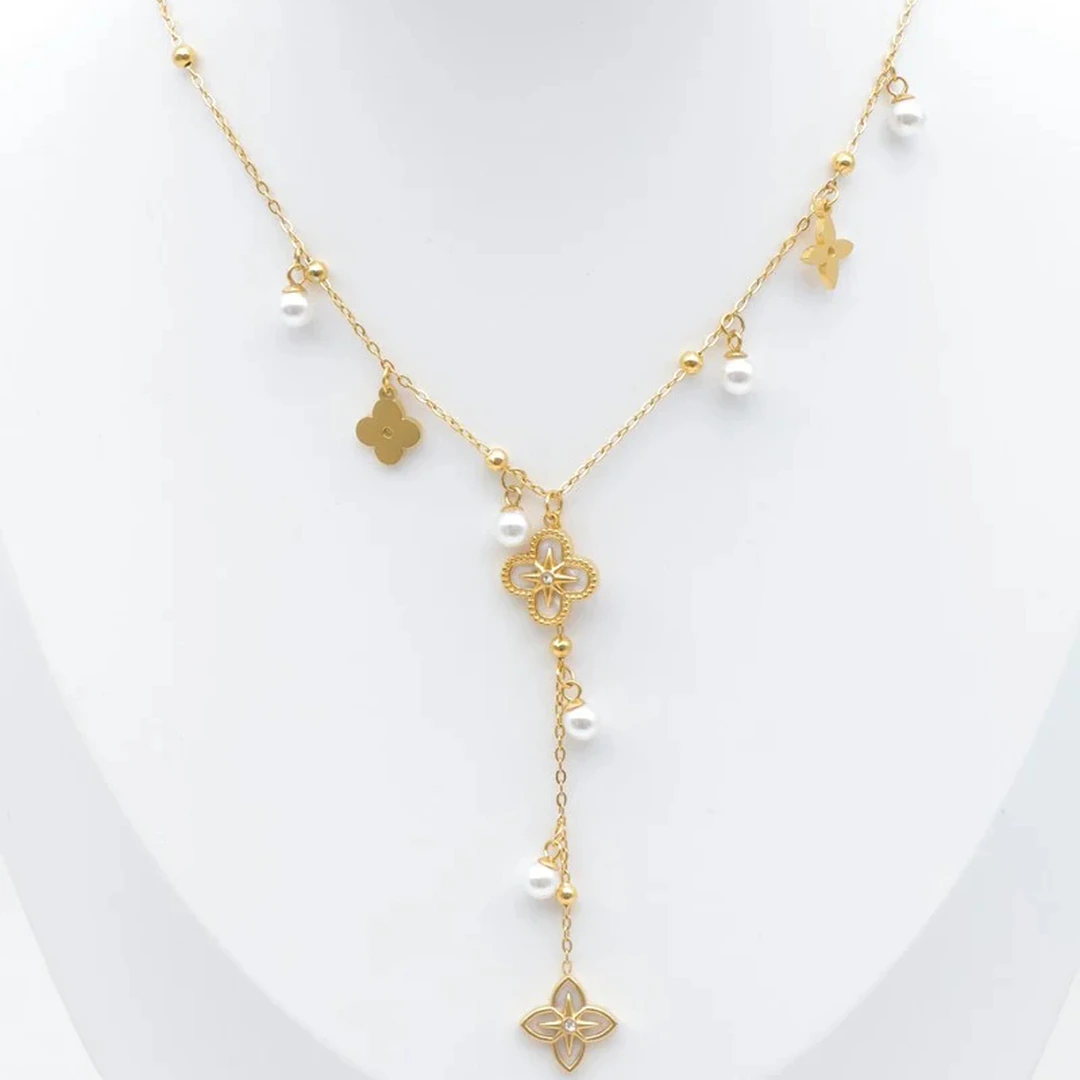 Collier doré à chute centrale, trèfle ajouré, petits charms dorés et perles blanches présenté sur un buste blanc