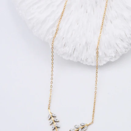 Collier doré femme à motif feuillage en V serti de pierres claires, posé près d'un décor blanc
