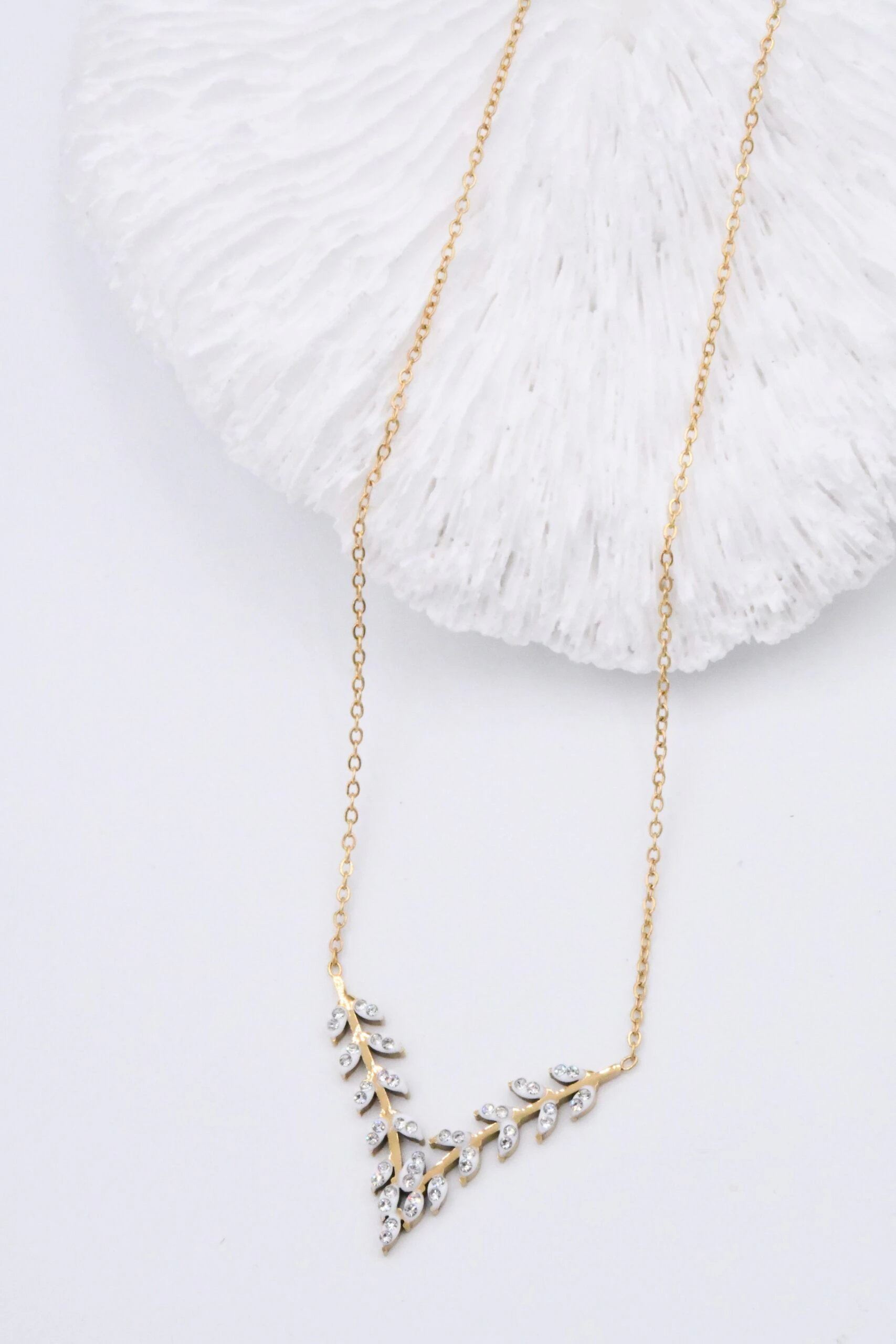 Collier doré femme à motif feuillage en V serti de pierres claires, posé près d'un décor blanc