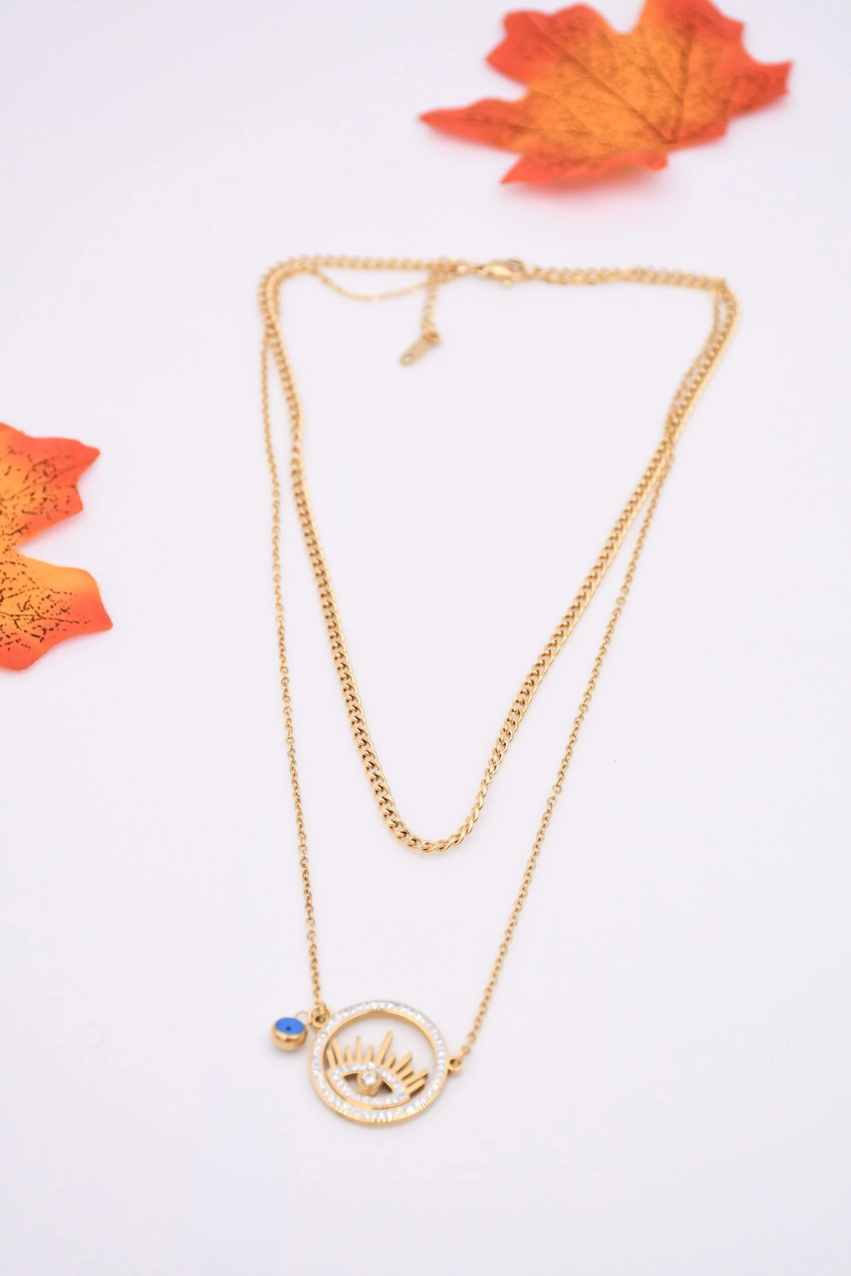 Collier doré femme double rang à pendentif mauvais œil ajouré serti de strass et breloque bleue, posé à plat