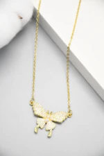 Collier papillon doré posé sur fond gris, détail des ailes pavées de strass et du corps perlé