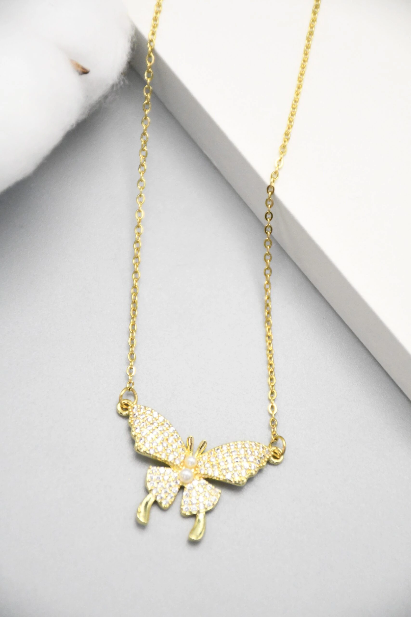 Collier papillon doré posé sur fond gris, détail des ailes pavées de strass et du corps perlé