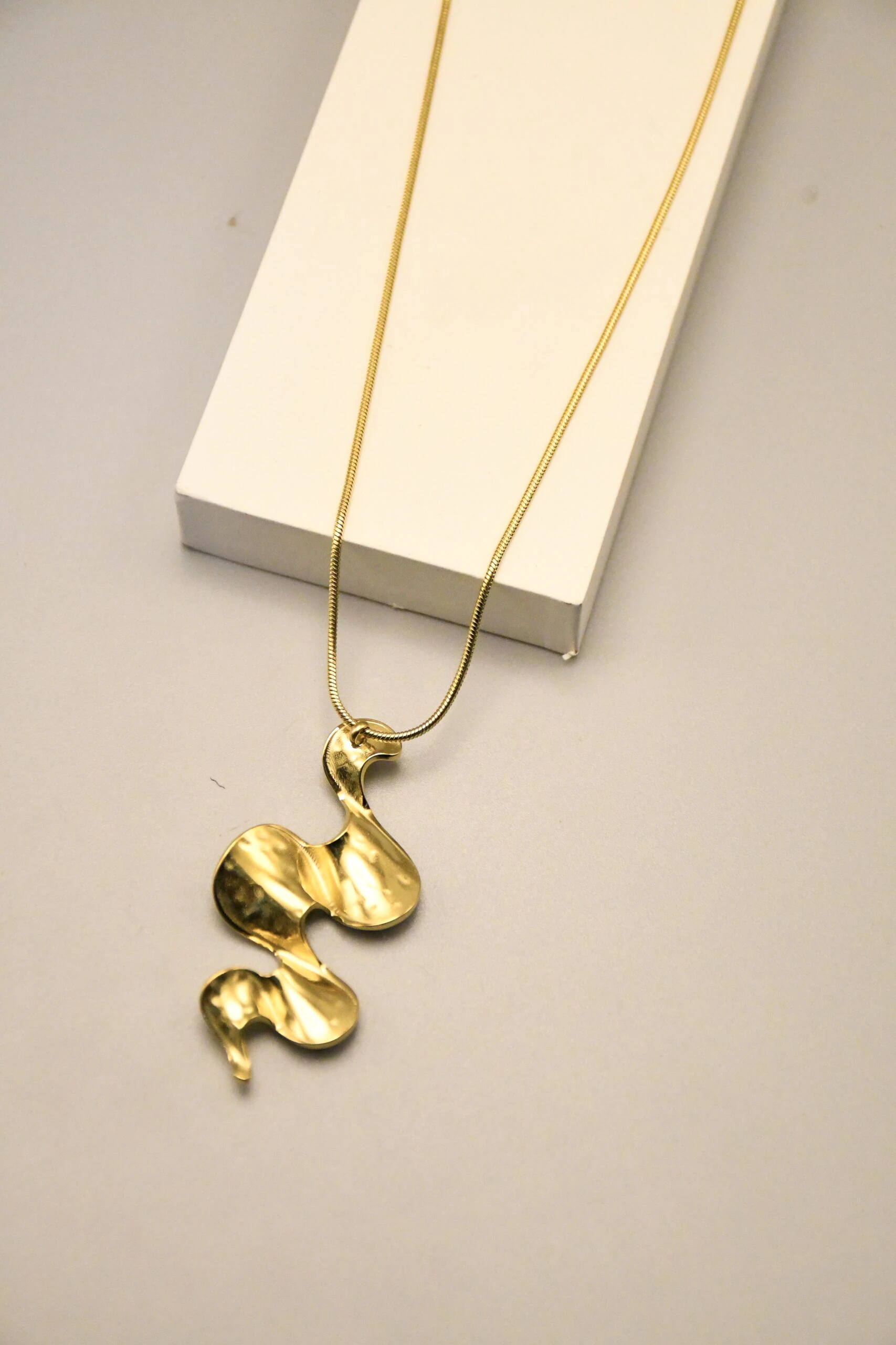 Collier à pendentif doré aux courbes ondulées posé contre un bloc blanc, vue rapprochée