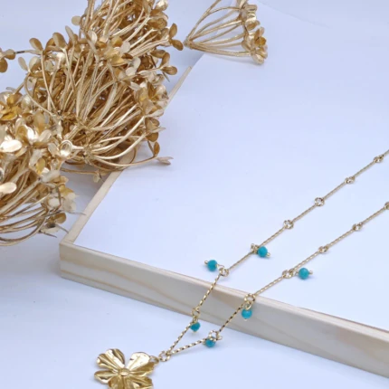 Collier chaîne fine dorée à perles bleu turquoise et fleur dorée posé près de fleurs séchées